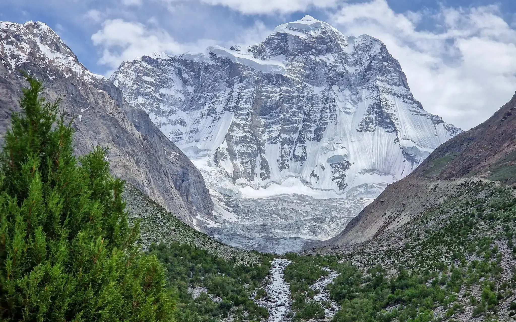 A glacier descending from a snow-covered peak, with its meltwater stream running down the valley below, in northern Pakistan.